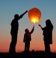 Silhuetter av tre personer som släpper iväg en tänd ljuslykta mot en färgglad himmel.