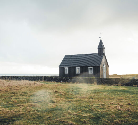En liten träkyrka står ensam på ett öppet fält under en molnig himmel.