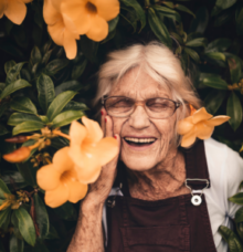 Äldre kvinna med glasögon står leende bland gula blommor.