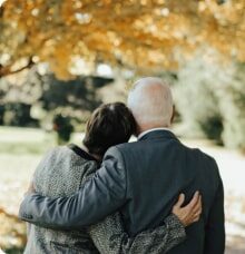 Två personer med grått hår omfamnar varandra under ett träd med gula löv.