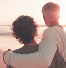 Två personer står omfamnade och tittar ut över havet vid solnedgången.