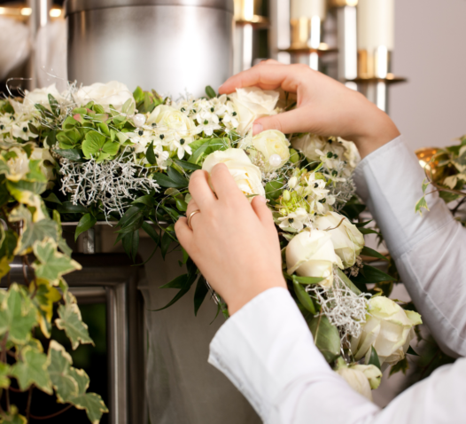 Händer arrangerar blommor och lövverk runt en metallbehållare med levande ljus i bakgrunden.