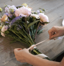Händer knyter ett band runt en bukett med blommor på ett träbord.