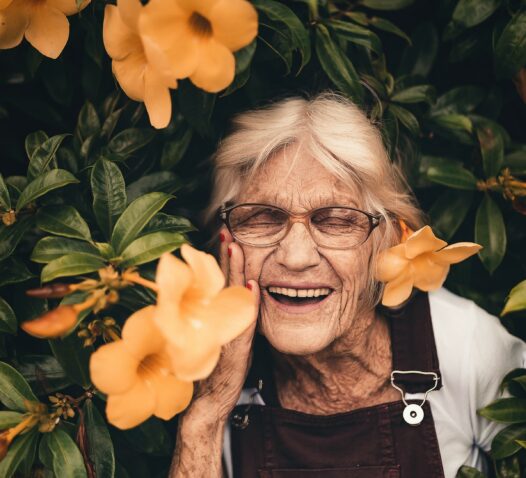 En äldre kvinna ler omgiven av orange blommor och gröna blad.