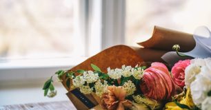 Blommor i olika färger och sorter ligger på ett bord framför ett fönster, inslagna i brunt papper.