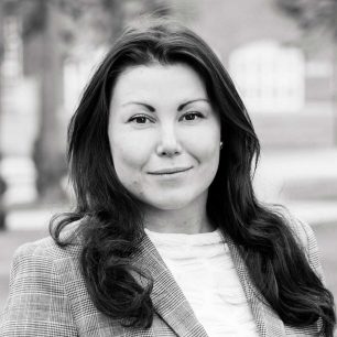 A woman with long hair wearing a plaid blazer and a white top, standing outdoors with blurred buildings in the background.