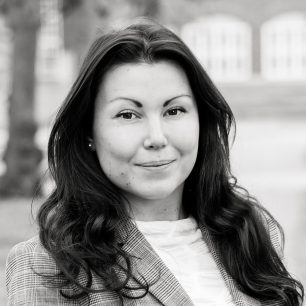 A black and white portrait of a person with long dark hair, wearing a checked blazer and a white top, smiling slightly while looking at the camera.