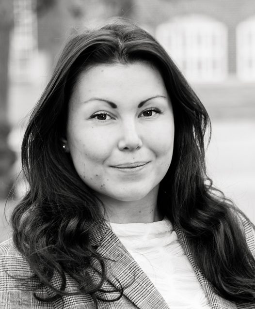 A black and white portrait of a person with long dark hair, wearing a checked blazer and a white top, smiling slightly while looking at the camera.