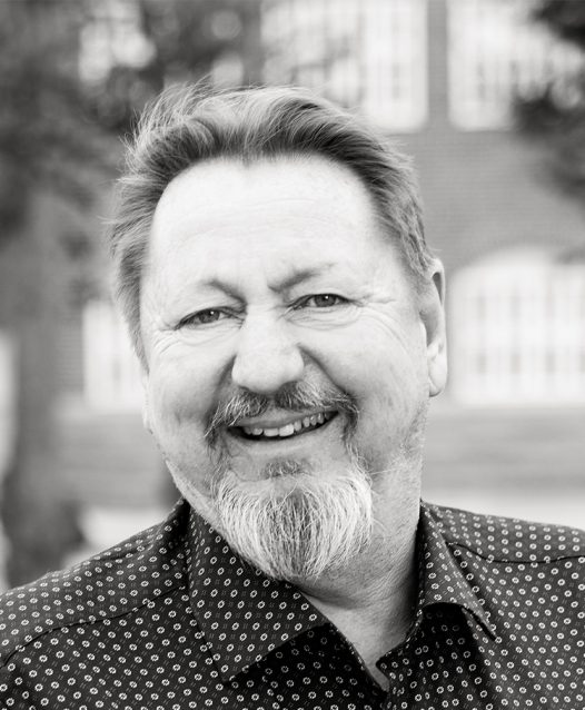 A person with a beard and mustache smiling, wearing a patterned shirt, with a blurred building in the background.